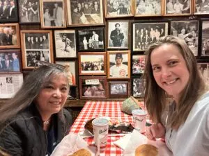 Two women seated at a table, smiling. A wall covered in photos of famous customers of the restaurant is in the background.
