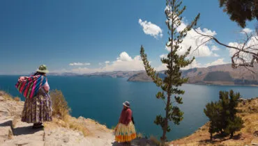 Two Women In Traditional Bolivian Clothes Standing On The Rock Close To The Titicaca Lake.